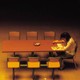 Yellow-tinted photograph of a woman reading while sitting alone at a table with six empty chairs next to her.